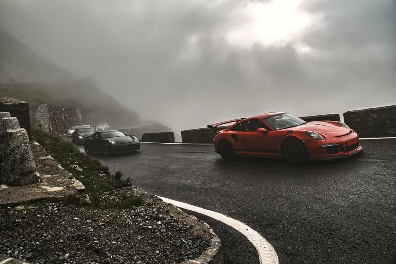 Porsche 911 GT3 RS in leuchtendem Orange auf nasser Passstraße im Nebel am Stilfser Joch (Stelvio), Sportwagen-Kolonne im Hintergrund